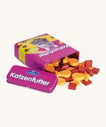 A open purple Erzi cat food tin lying flat next to the lid with the different shaped cat food wooden pieces pouring out of the tin.