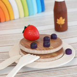 Erzi wooden toy breakfast scene with stacked round pancakes on a plate, a red strawberry, purple blueberries, a small brown bottle with a maple leaf, and wooden utensils on a striped surface.