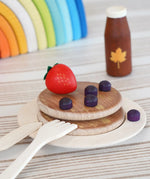 Erzi wooden toy breakfast scene with stacked round pancakes on a plate, a red strawberry, purple blueberries, a small brown bottle with a maple leaf, and wooden utensils on a striped surface.
