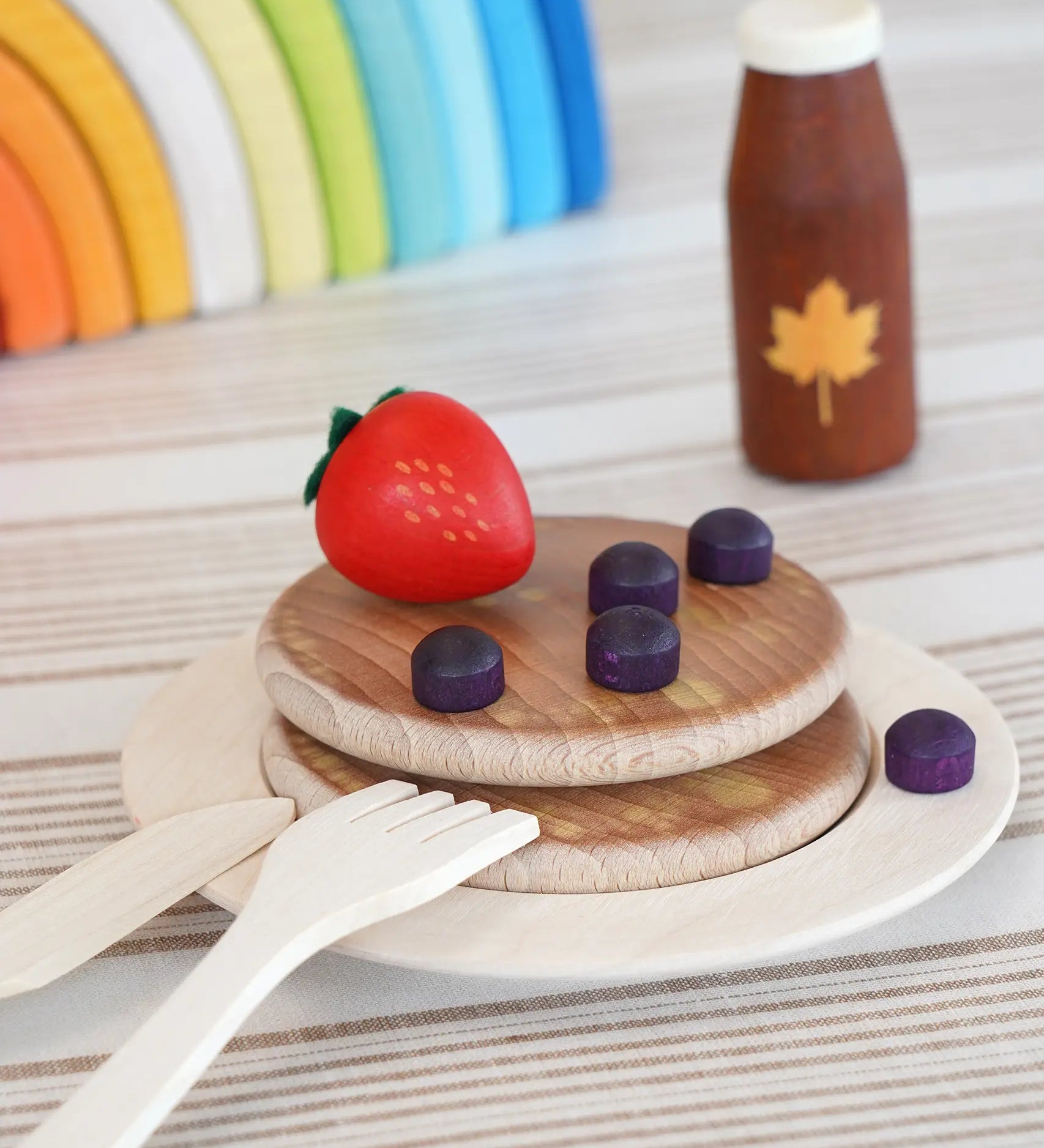 Erzi wooden toy breakfast scene with stacked round pancakes on a plate, a red strawberry, purple blueberries, a small brown bottle with a maple leaf, and wooden utensils on a striped surface.