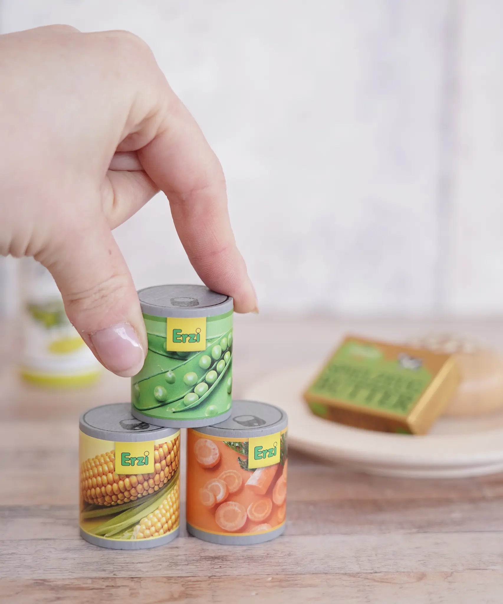 Erzi Tinned Vegetables (carrots, peas, sweetcorn), wooden canned veggie play food, stacked in a play set up with a hand for scale.