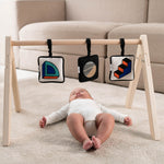 A baby lying down on their back, underneath a wooden baby gym, with the Etta Loves x Camille Walala Sensory Hanging Squares attached to the rung across the top of the baby gym. The baby is looking upwards towards the sensory squares, looking at the shapes and prints