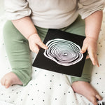 A baby playing with the Etta Loves Sensory Positive Affirmation Flash Cards. The card being held shows a black and white spiral 