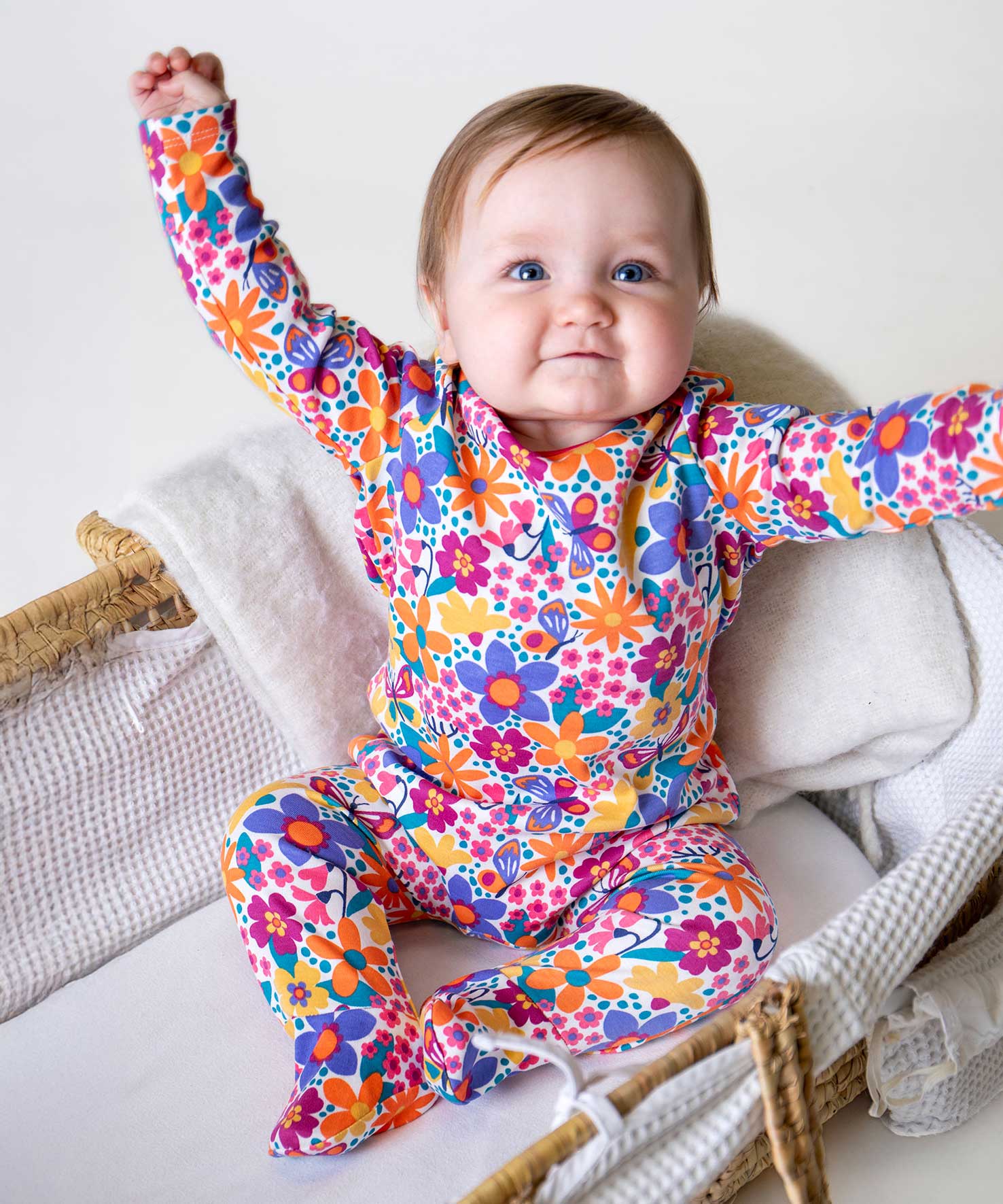 A baby sitting up wearing the Frugi easy dressing printed babygrow in white with aretro floral design. A GOTS organic cotton all in one with a bright retro floral design and pink trim around the neck and shoulder. This babygrow is a part of a wide range of organic cotton clothing for children available here at Babipur. 