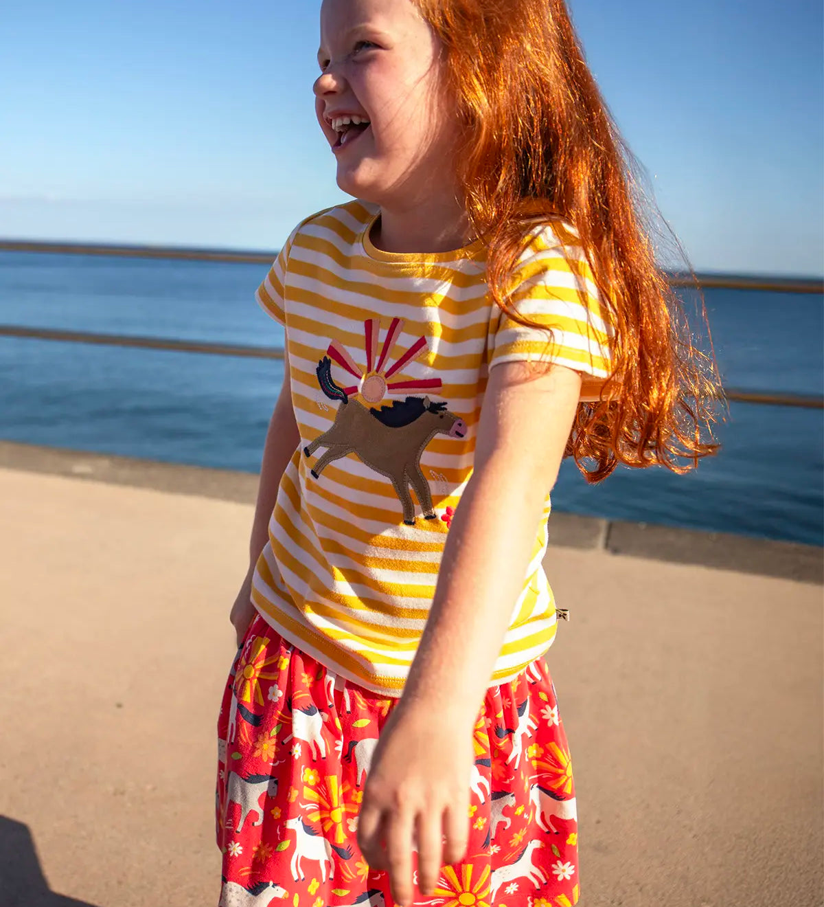 Small child wearing Frugi 100% organic cotton short sleeve t-shirt in yellow and white stripe with jumping horse patch with the ocean in the background.