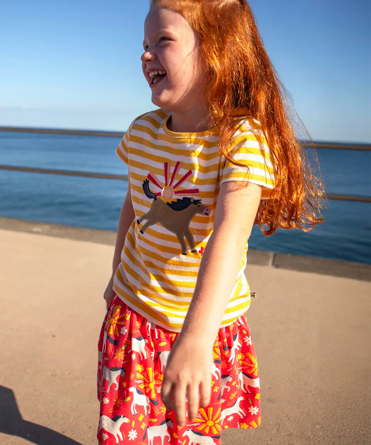 Small child wearing Frugi 100% organic cotton short sleeve t-shirt in yellow and white stripe with jumping horse patch with the ocean in the background.