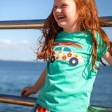 Child waering Frugi 100% organic cotton short sleeve t-shirt in light blue with white car patch on the front. Child standing against a metal railing