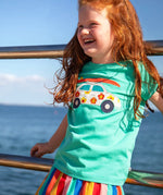 Child waering Frugi 100% organic cotton short sleeve t-shirt in light blue with white car patch on the front. Child standing against a metal railing