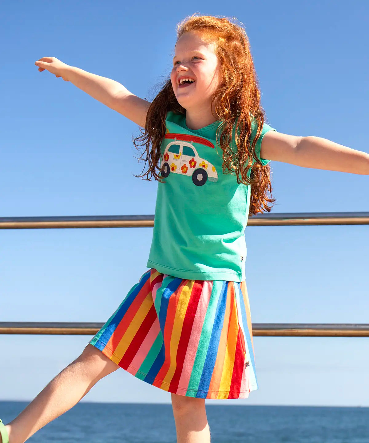Child wearing Frugi Alba skort in rainbow stripes with the ocean in the background