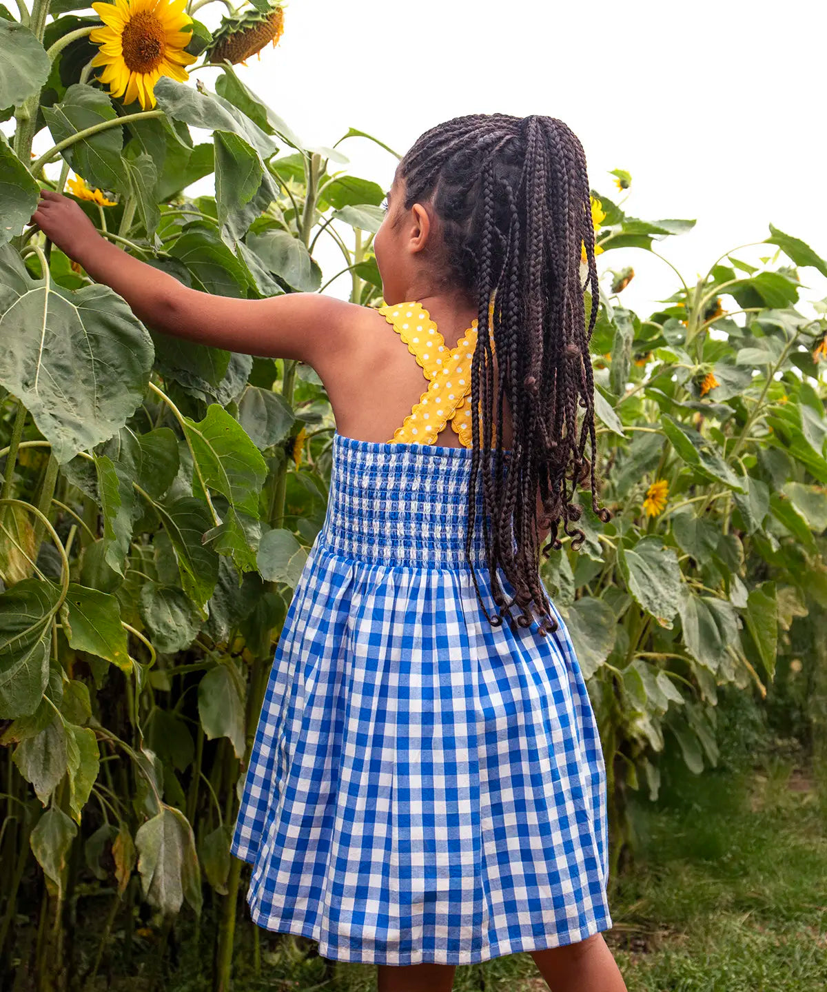 Back of child wearing Frugi 100% organic cotton dress in blue and white gingham showing yellow shoulder straps and elasticated upper body