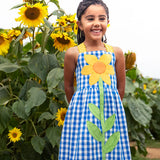 Child wearing Frugi 100% organic cotton dress in blue and white gingham with a large sunflower patch on front in a sunflower field