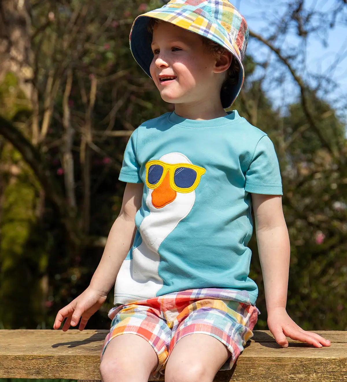 Child wearing Frugi's Frugi 100% organic cotton Ashley reversible bucket hat in pastel check and sitting on a wooden surface