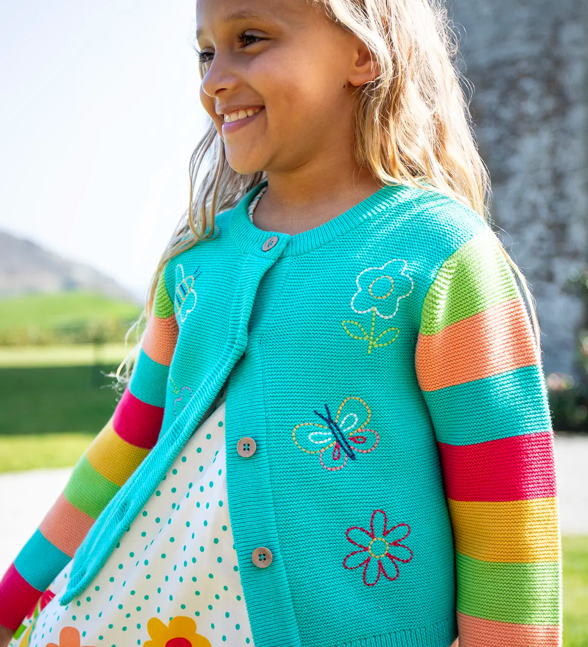 Child wearing Frugi 100% organic cotton cammie cardigan in light blue with rainbow striped sleeves showing spring embroideries on the front