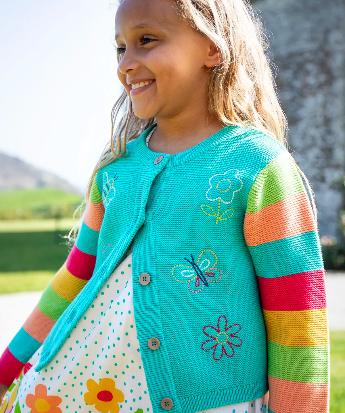 Child wearing Frugi 100% organic cotton cammie cardigan in light blue with rainbow striped sleeves showing spring embroideries on the front