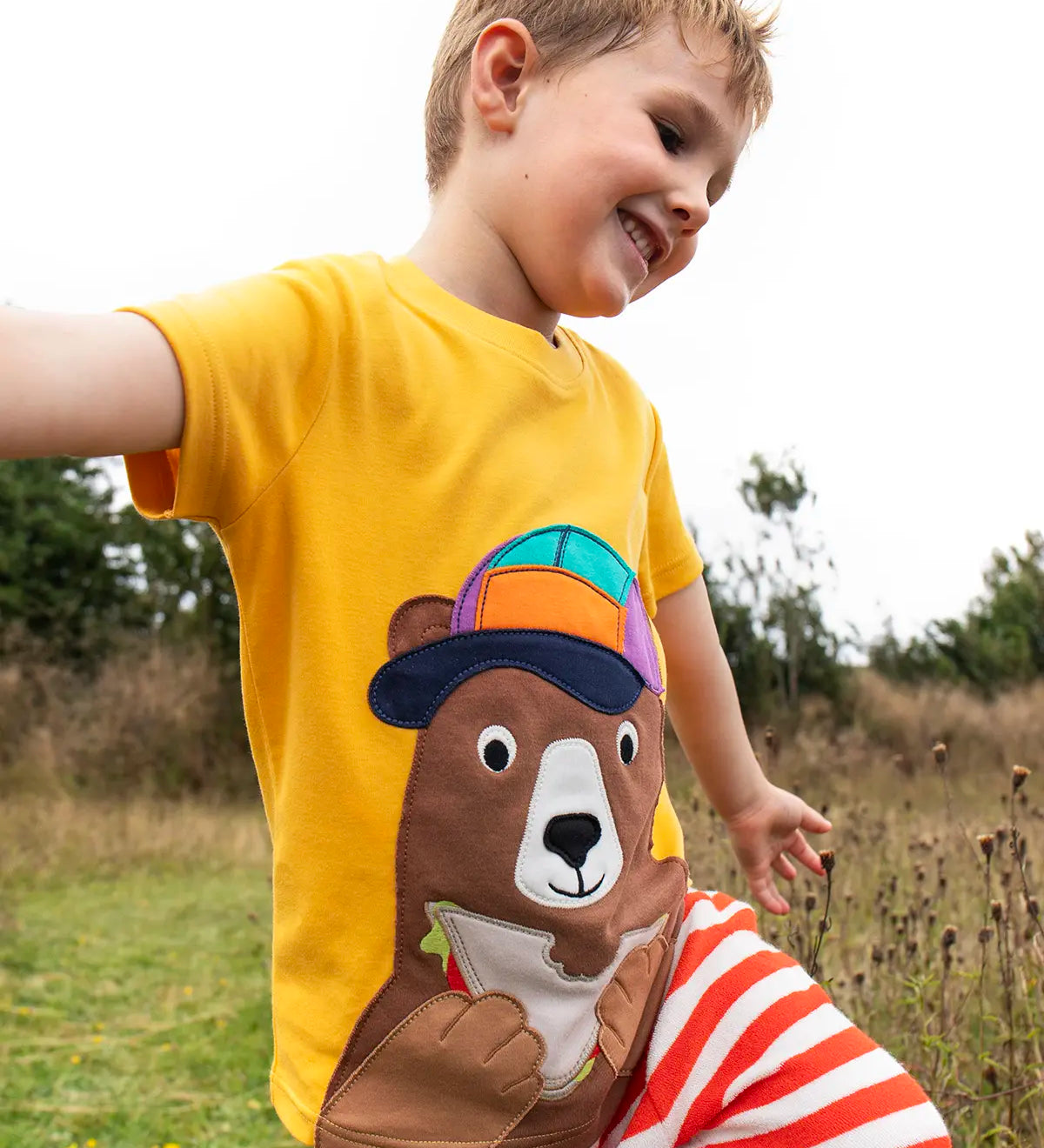 Child wearing the Frugi 100% organic cotton yellow short sleeve t-shirt with a playful brown bear eating a sandwich patch in a grassy field