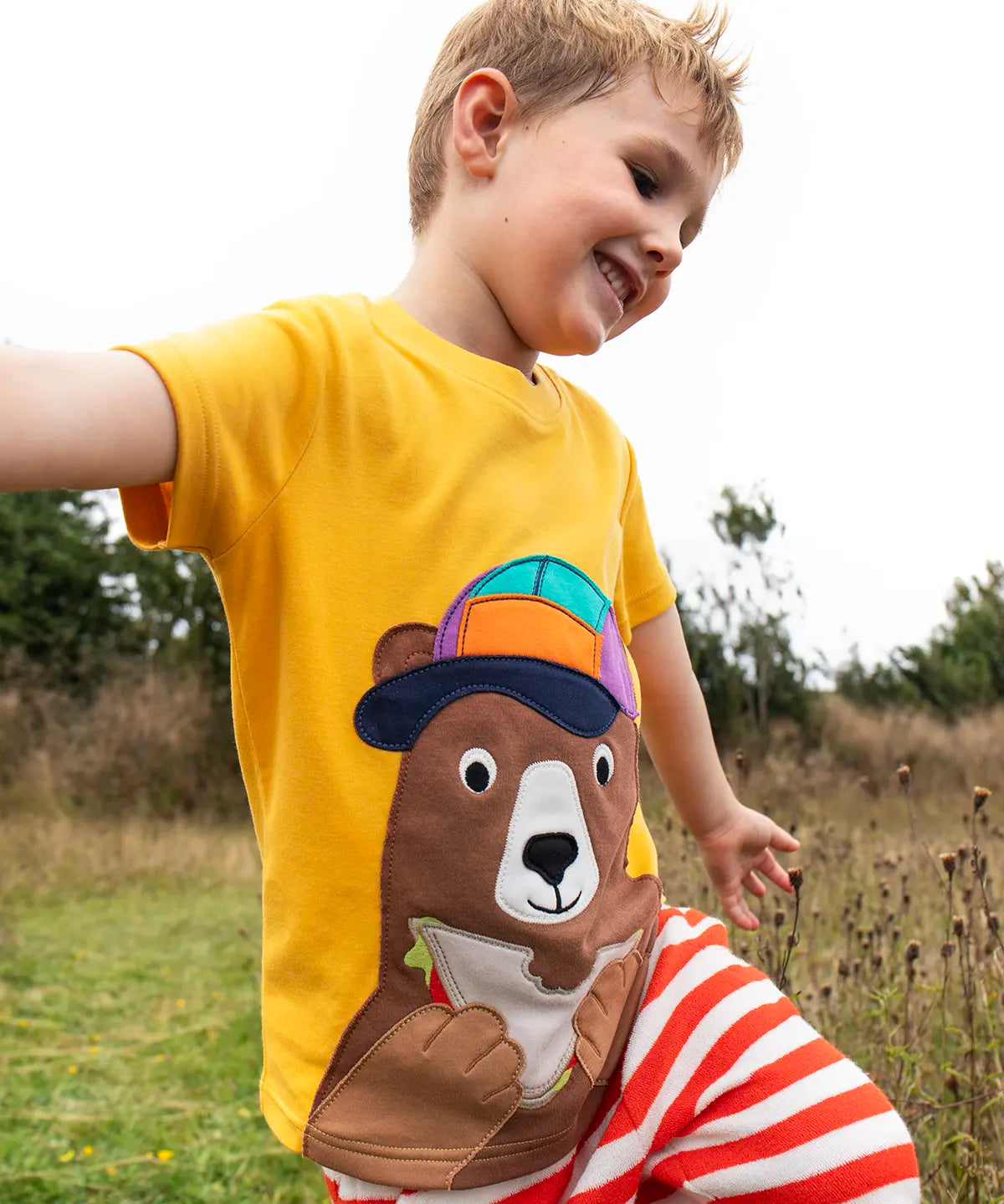 Child wearing the Frugi 100% organic cotton yellow short sleeve t-shirt with a playful brown bear eating a sandwich patch in a grassy field