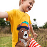 Child wearing the Frugi 100% organic cotton yellow short sleeve t-shirt with a playful brown bear eating a sandwich patch in a grassy field