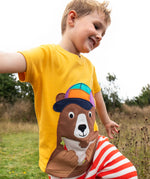 Child wearing the Frugi 100% organic cotton yellow short sleeve t-shirt with a playful brown bear eating a sandwich patch in a grassy field