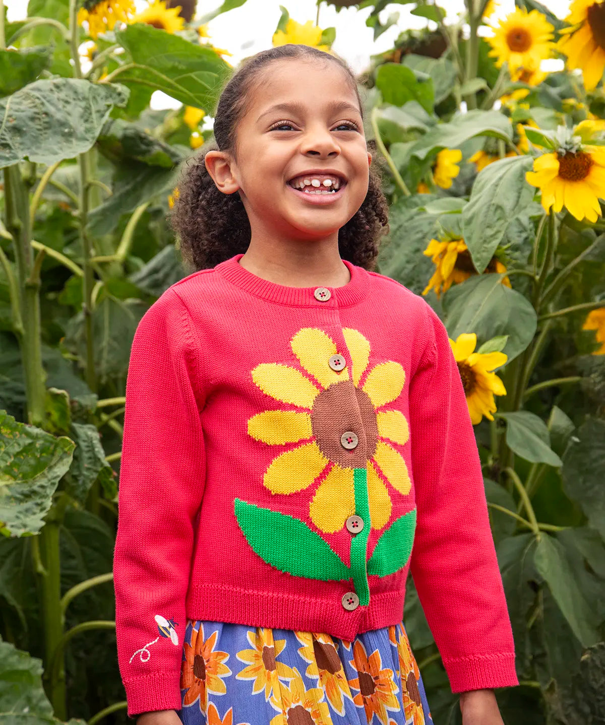 Child wearing Frugi 100% organic cotton Ella cardigan in pink with sunflower design on the front with tall sunflowers in the background