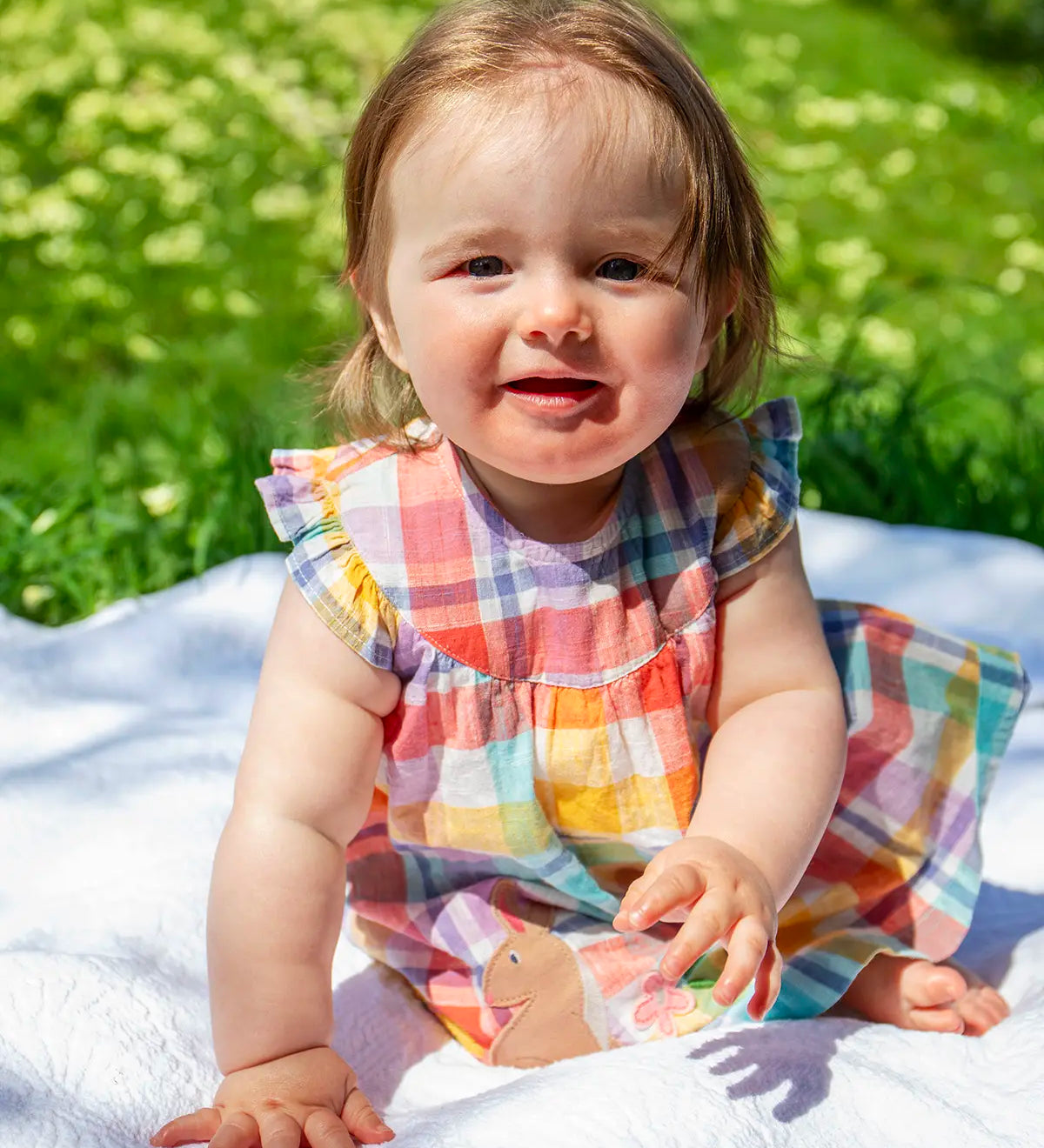 Child wearing Frugi Flora sleeveless dress in a pastel check design with a rabbit patch on the skirt and sitting on a blanket