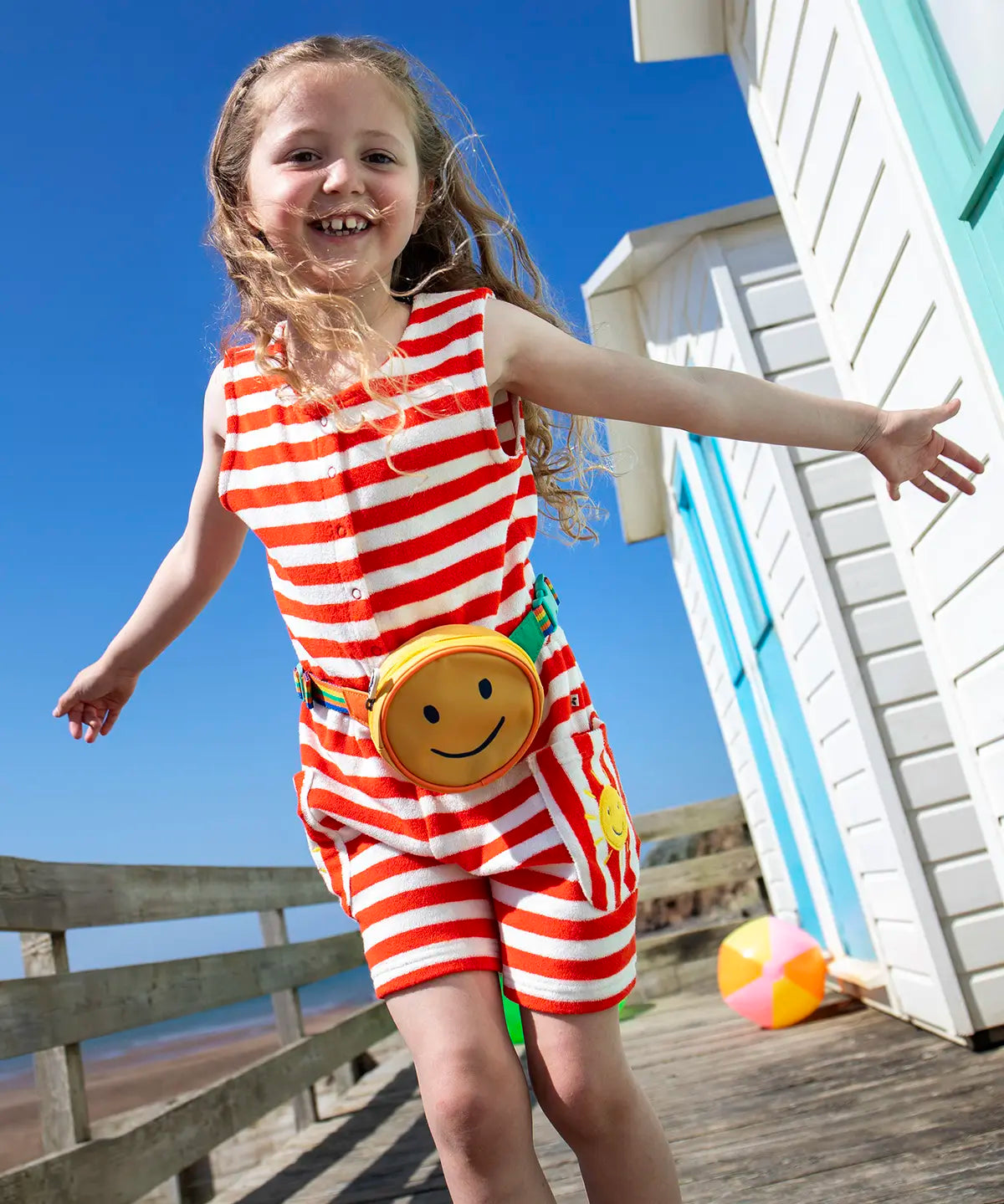 Child wearing Frugi 100% organic cotton Harlyn towelling playsuit in red and white stripes on a wooden surface