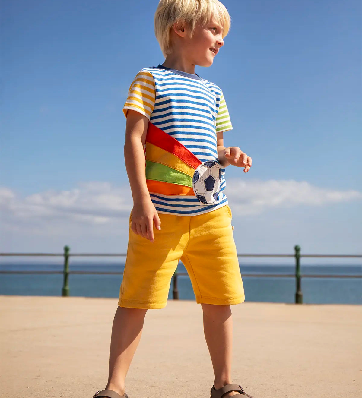 Child wearing Frugi's 100% organic cotton striped short sleeve t-shirt with a football patch and yellow shorts. Child standing on a concrete surface