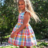 Side of child waering Frugi springtime dress in pastel check with hyacinth embroideries on the upper body with trees in the background