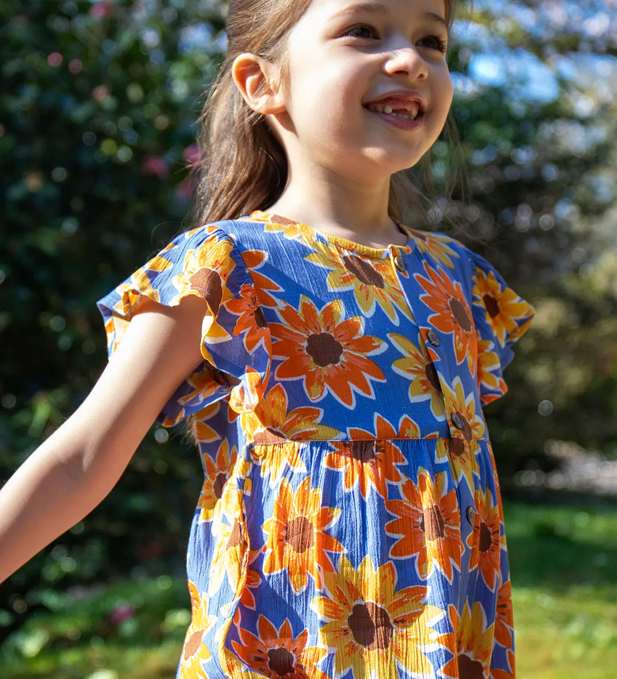 Child wearing Frugi Lowenna jumpsuit in purple with a repeating sunflower print and frilled shoulder with trees in the background.