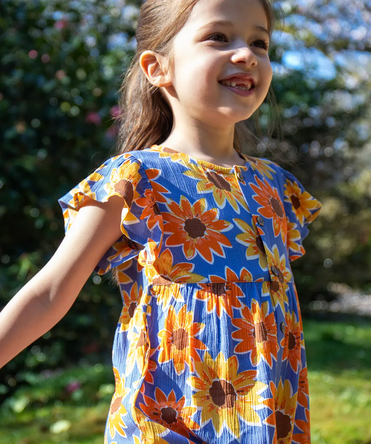 Child wearing Frugi Lowenna jumpsuit in purple with a repeating sunflower print and frilled shoulder with trees in the background.