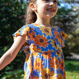 Child wearing Frugi Lowenna jumpsuit in purple with a repeating sunflower print and frilled shoulder with trees in the background.