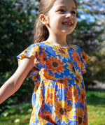 Child wearing Frugi Lowenna jumpsuit in purple with a repeating sunflower print and frilled shoulder with trees in the background.