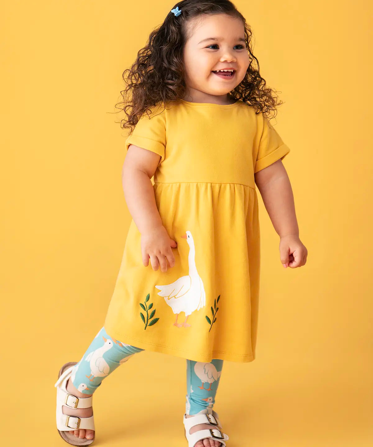 Child wearing Frugi's bumblebee goose gaggle outfit showing a yellow dress and blue leggings with a repeating goose print against a orange background