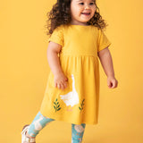 Child wearing Frugi's bumblebee goose gaggle outfit showing a yellow dress and blue leggings with a repeating goose print against a orange background