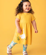 Child wearing Frugi's bumblebee goose gaggle outfit showing a yellow dress and blue leggings with a repeating goose print against a orange background
