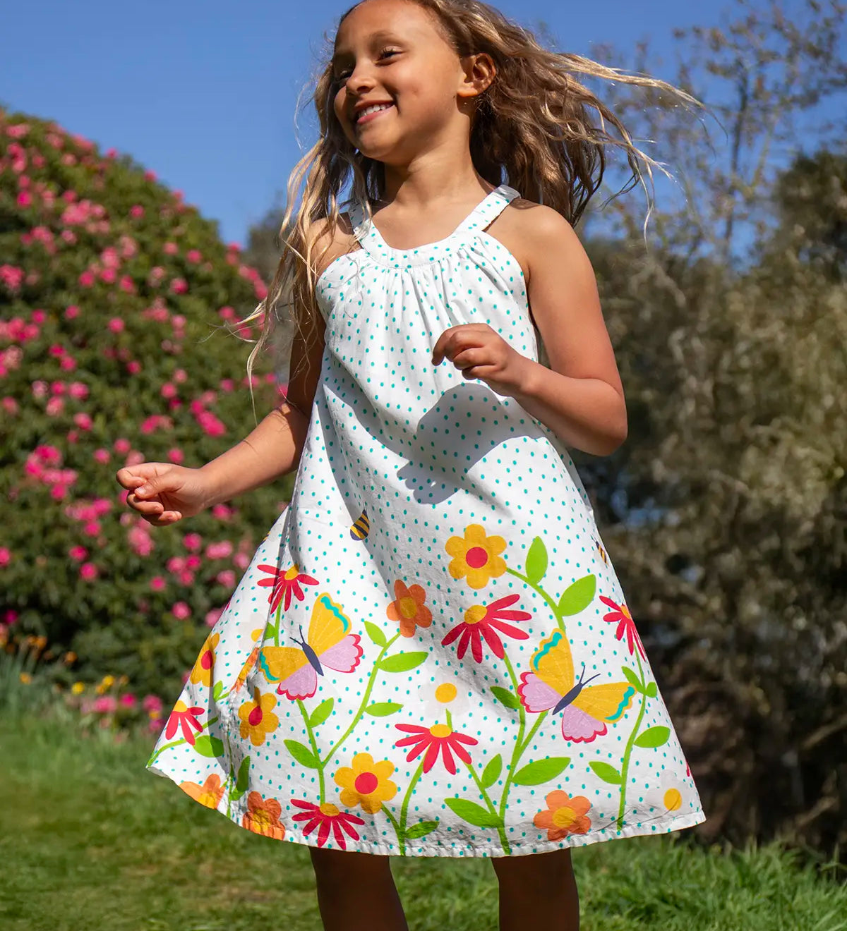 Child wearing Frugi 100% organic cotton Myra twirly dress in white with a repeating floral, bee and butterfly print. Child in a grassy field