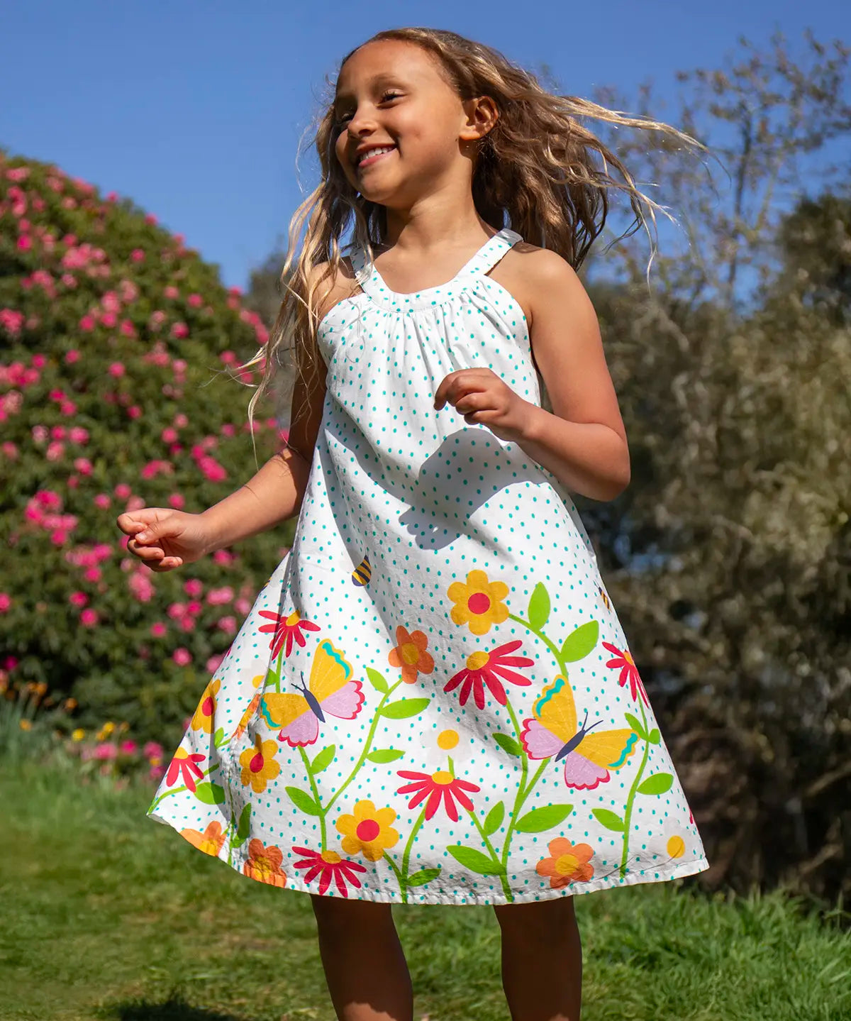Child wearing Frugi 100% organic cotton Myra twirly dress in white with a repeating floral, bee and butterfly print. Child in a grassy field