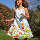 Child wearing Frugi 100% organic cotton Myra twirly dress in white with a repeating floral, bee and butterfly print. Child in a grassy field