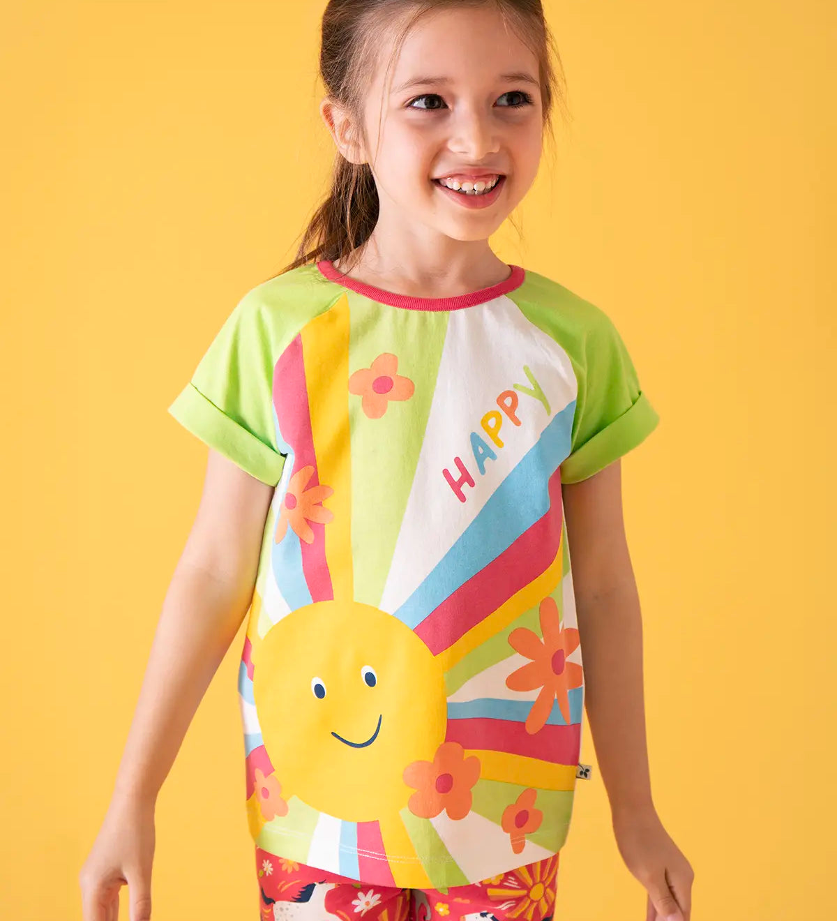 Child wearing Frugi 100% organic cotton light green raglan short sleeve t-shirt with a large happy sun print on the front against a orange background.