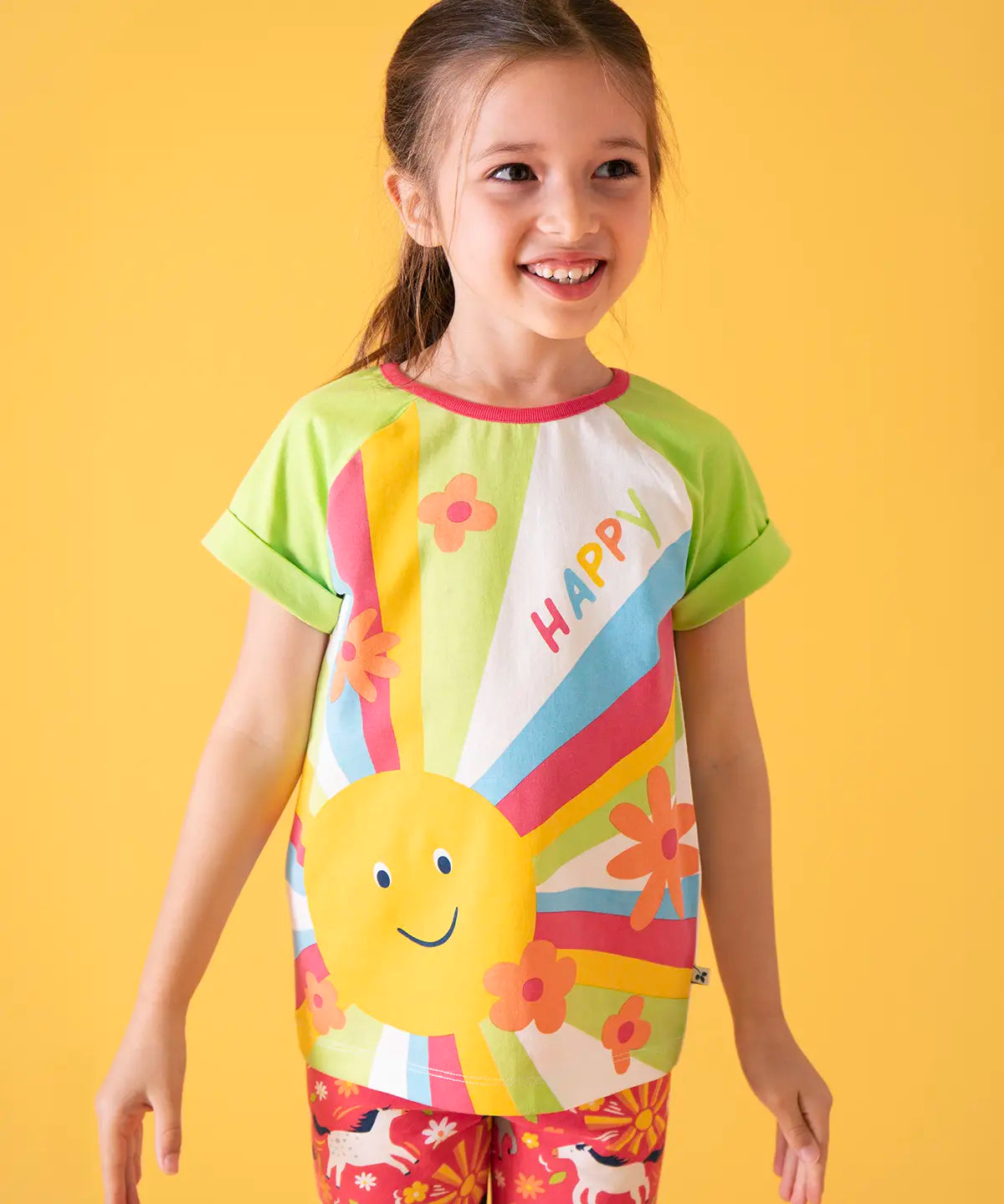 Child wearing Frugi 100% organic cotton light green raglan short sleeve t-shirt with a large happy sun print on the front against a orange background.