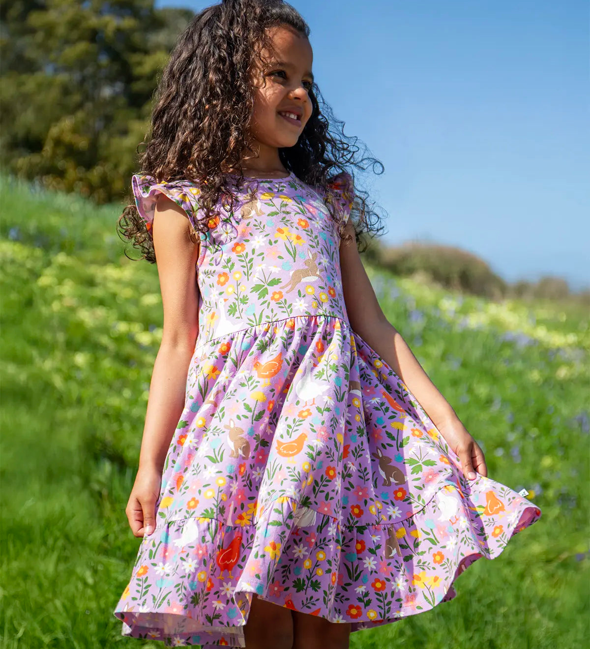 Child wearing Frugi's 100% organic cotton skater dress in violet purple with a repeating meadow inspired print on a grassy hill 
