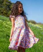 Child wearing Frugi's 100% organic cotton skater dress in violet purple with a repeating meadow inspired print on a grassy hill 