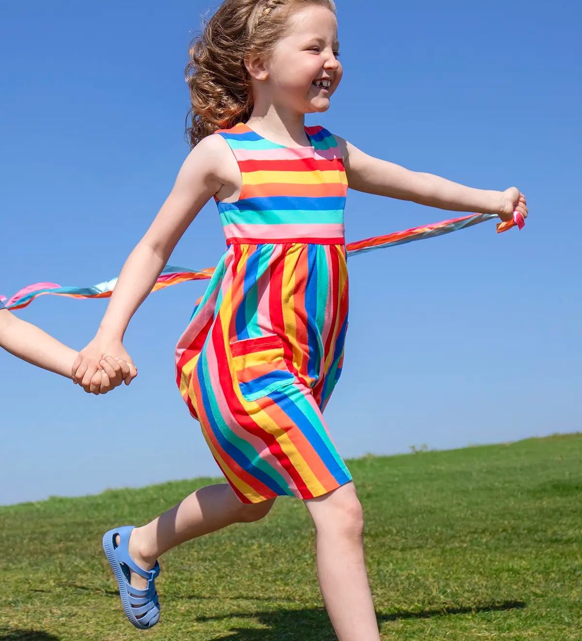 Child wearing Frugi 100% organic cotton rainbow striped Samantha Summer dress on a grassy field
