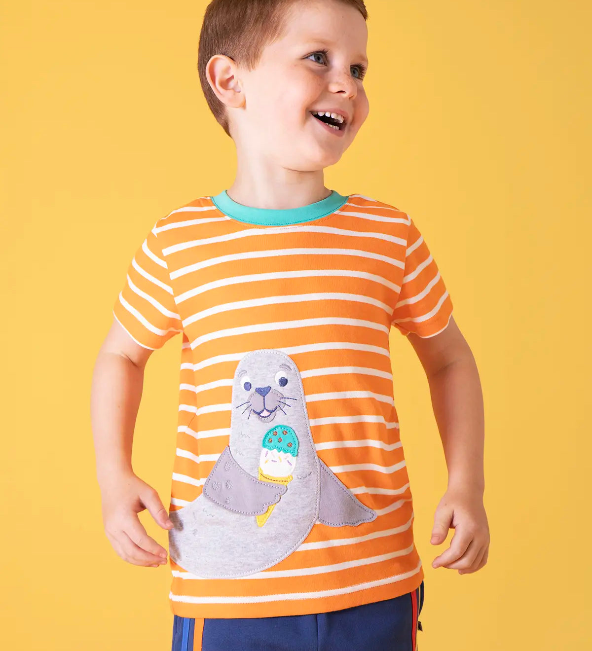 Child wearing Frugi 100% organic cotton short sleeve t-shirt in orange and white stripes with a seal patch on the front against a orange background