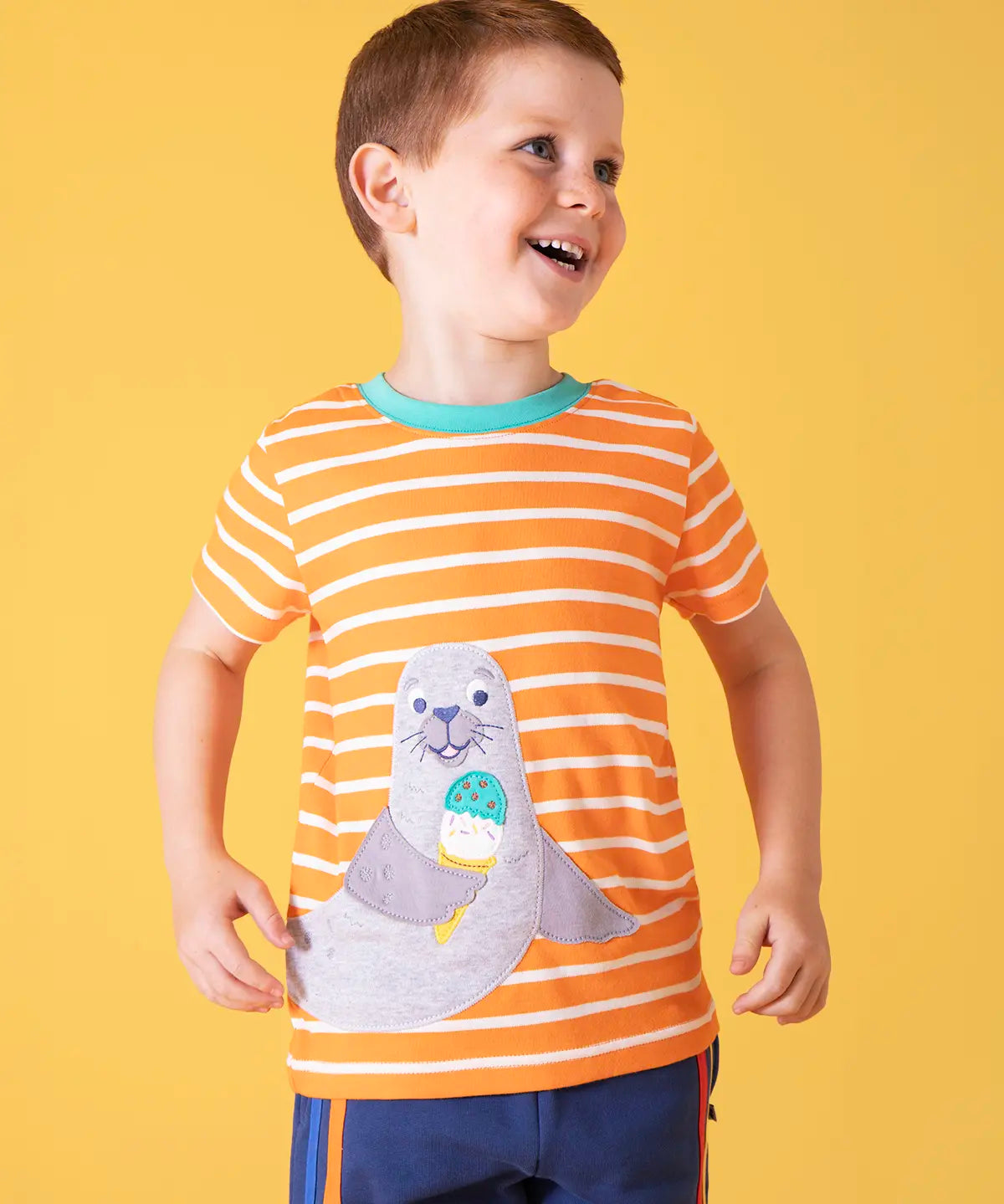 Child wearing Frugi 100% organic cotton short sleeve t-shirt in orange and white stripes with a seal patch on the front against a orange background