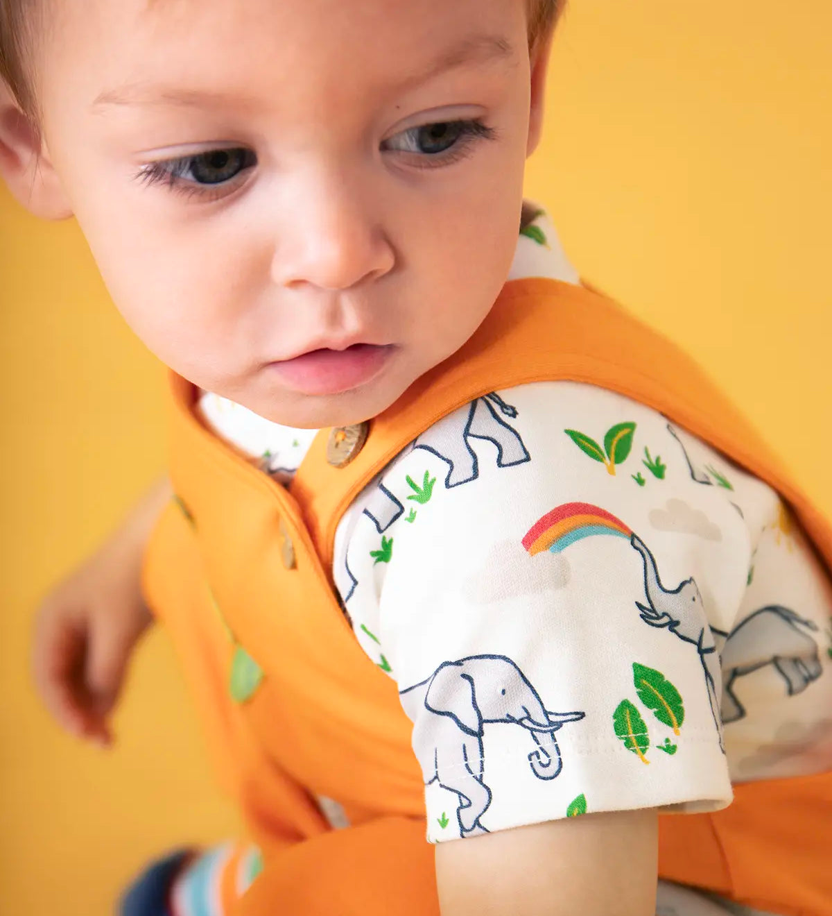 Baby wearing Frugi's 100% organic cotton body suit in white with a repeating elephant print against a orange background