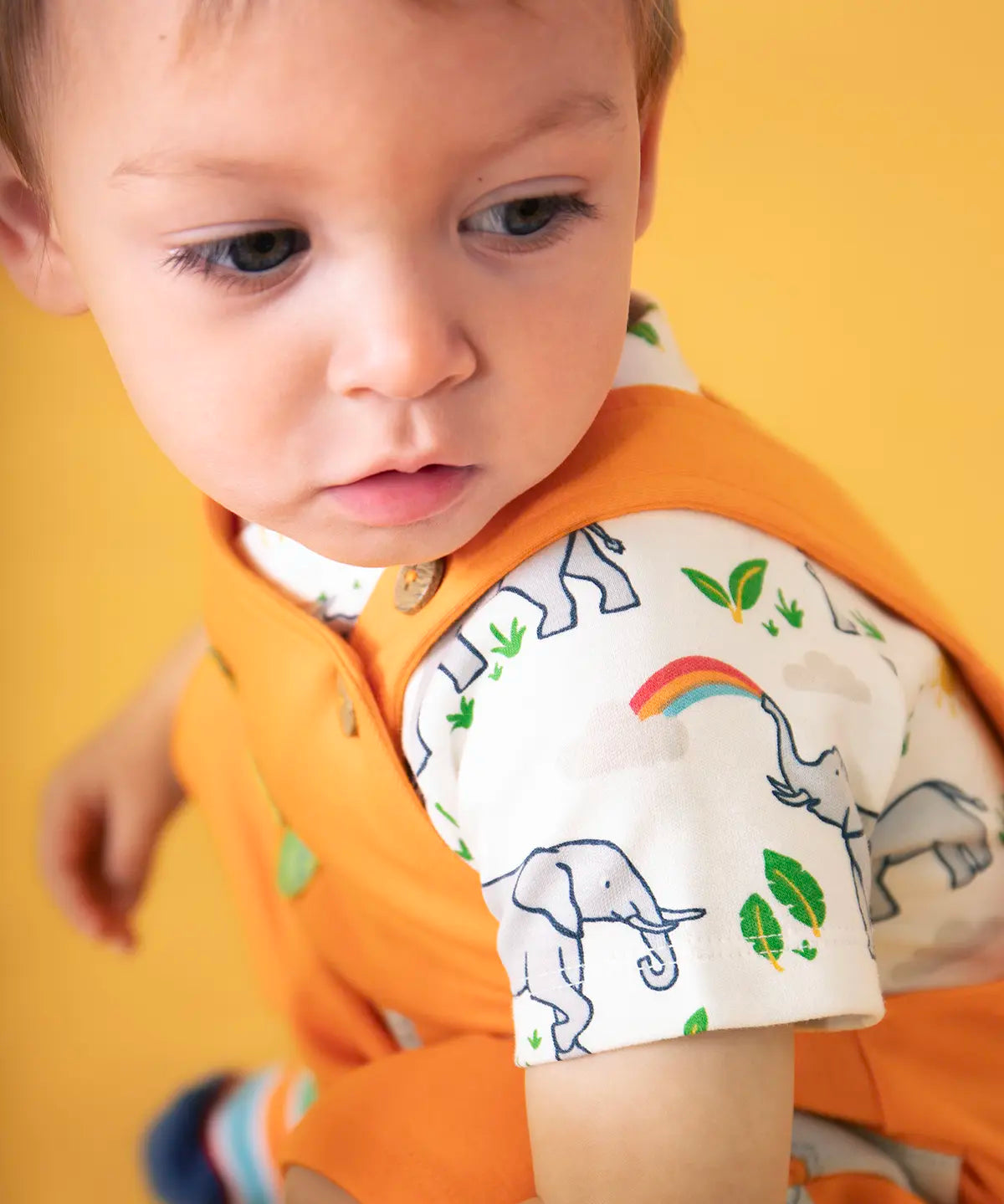 Baby wearing Frugi's 100% organic cotton body suit in white with a repeating elephant print against a orange background