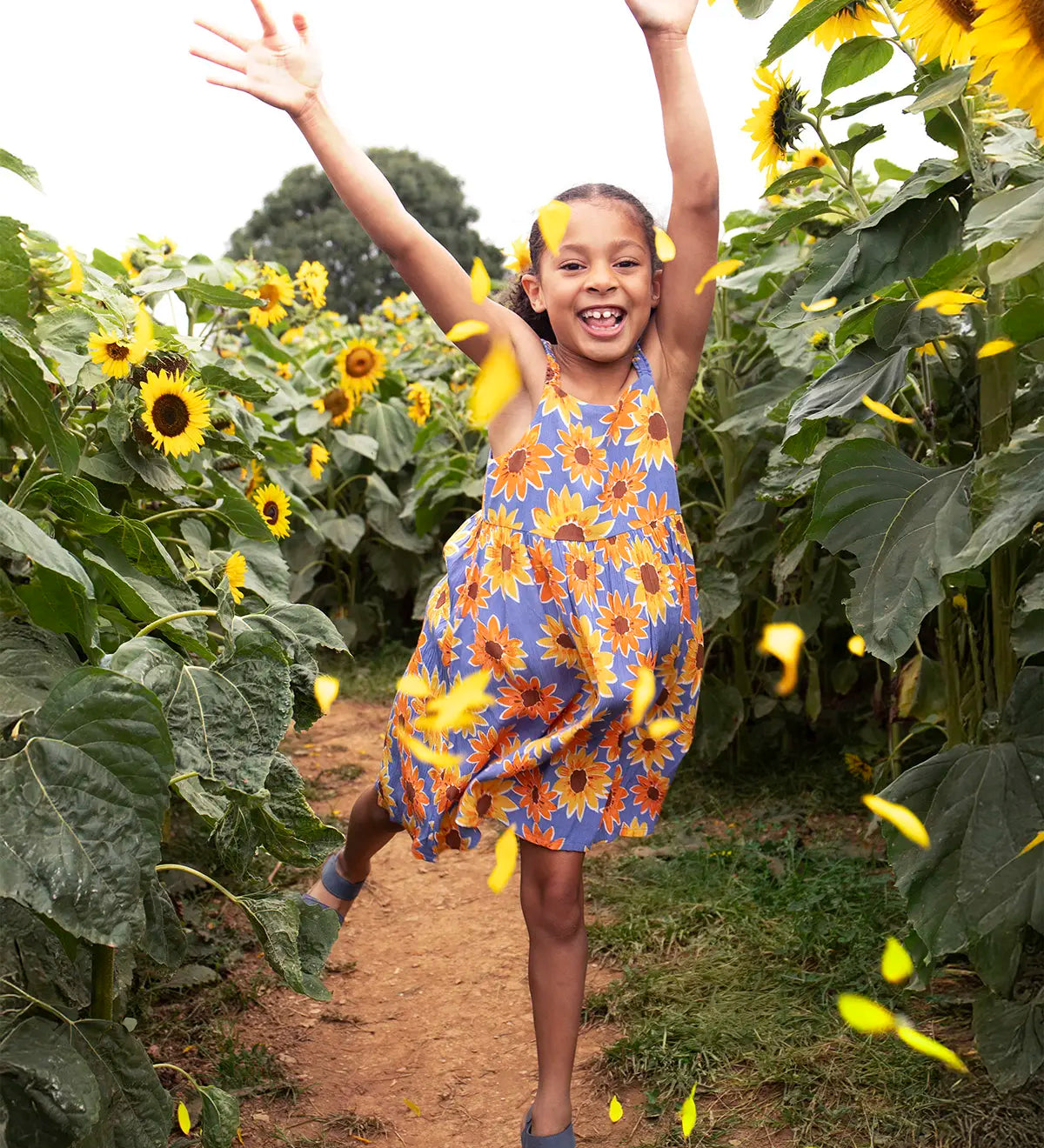 Child wearing Frugi 100% organic cotton Sophie summer sleeveless dress in purple with a repeating sunflower print in a sunflower field