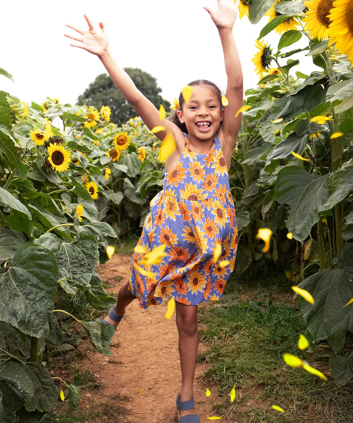 Child wearing Frugi 100% organic cotton Sophie summer sleeveless dress in purple with a repeating sunflower print in a sunflower field