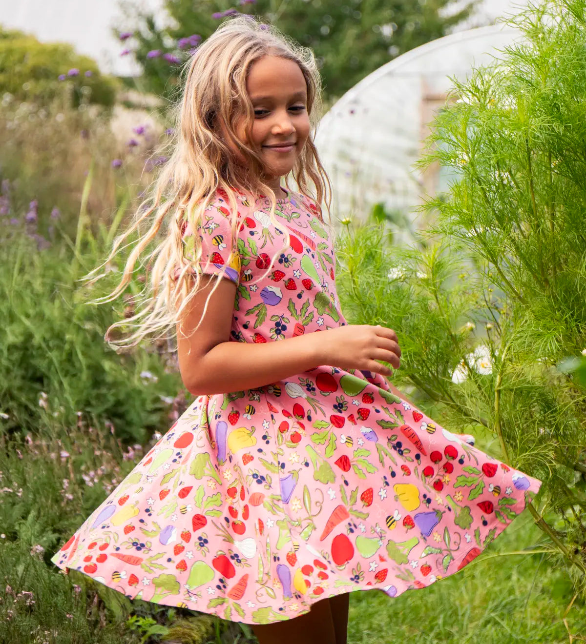 Child wearing Frugi's 100% organic cotton spring skater dress in pink with a repeating fruit and veg print with grass and flowers in the background