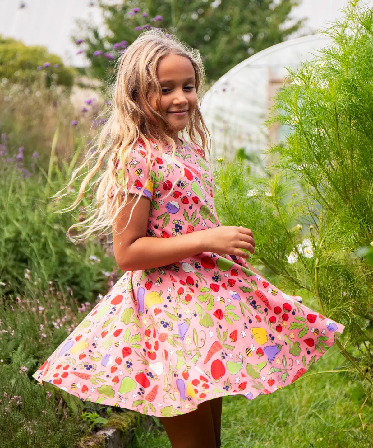 Child wearing Frugi's 100% organic cotton spring skater dress in pink with a repeating fruit and veg print with grass and flowers in the background